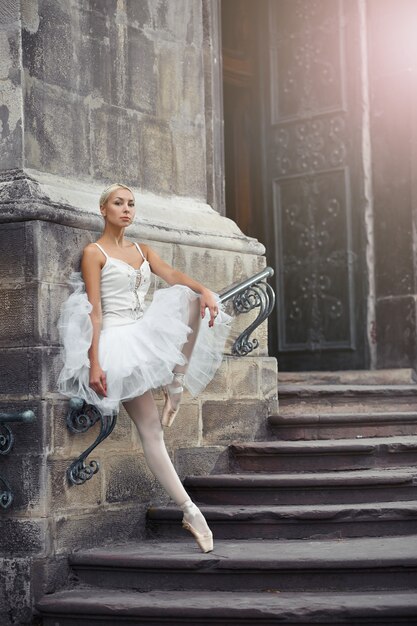 Portret van een mooie jonge blonde ballerina in witte outfit die sierlijk op de trappen van een oud kasteel staat.