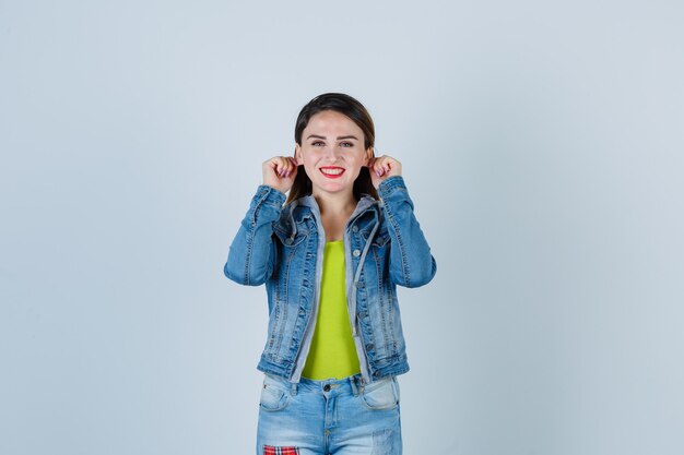 Portret van een mooie jonge dame die haar oorlellen in een spijkerbroek naar beneden trekt en er geamuseerd vooraanzicht uitziet