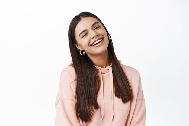 Portret van een mooie jonge openhartige vrouw knipoogt naar voren, glimlacht en lacht, staat gelukkig in hoodie tegen een witte muur, blijft positief