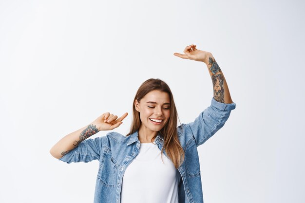 Portret van een mooie jonge vrouw die plezier heeft, gelukkig lacht en de handen vrij steekt, zorgeloos danst, in vrijetijdskleding op wit staat