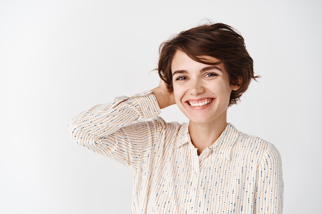 Portret van een mooie jonge vrouw met een witte glimlach, die in een blouse staat en kort haar aanraakt, die over een witte muur staat