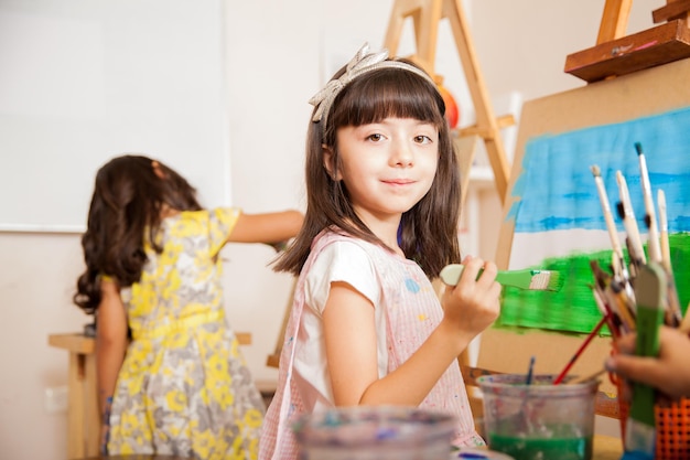 Portret van een mooie kleine kunstenaar die een penseel vasthoudt en glimlacht terwijl hij aan een schilderij werkt voor de kunstles