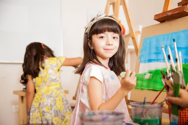 Portret van een mooie kleine kunstenaar die een penseel vasthoudt en glimlacht terwijl hij aan een schilderij werkt voor de kunstles
