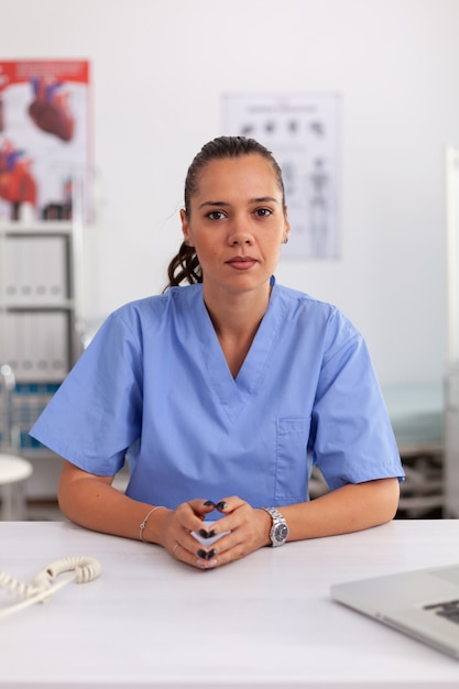 Portret van een mooie medische verpleegster die lacht naar de camera in het ziekenhuiskantoor met een blauw uniform. Gezondheidszorgbeoefenaar zit aan bureau met behulp van computer in moderne kliniek die naar monitor, geneeskunde kijkt.