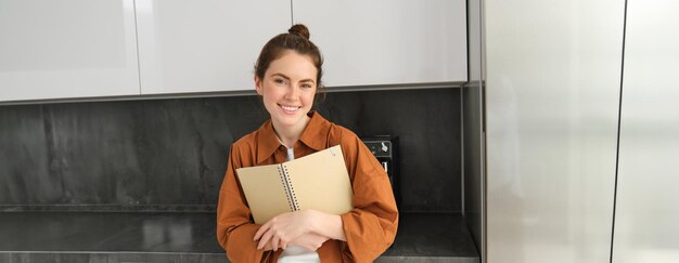 Portret van een mooie moderne vrouw die in de keuken staat met een receptenboek en een notitieboek.