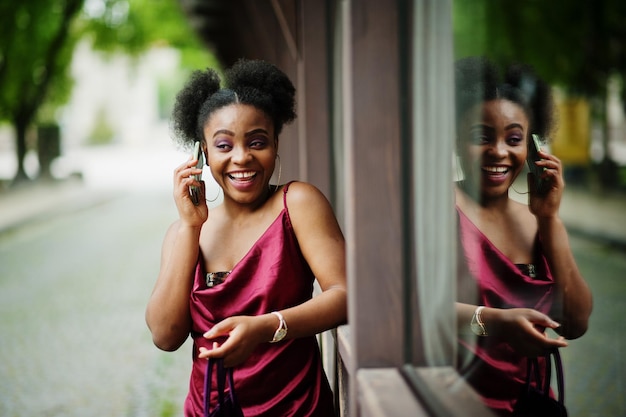 Portret van een mooie natuurlijke jonge Afrikaanse vrouw met afrohaar Zwart model in rode zijden jurk met mobiele telefoon