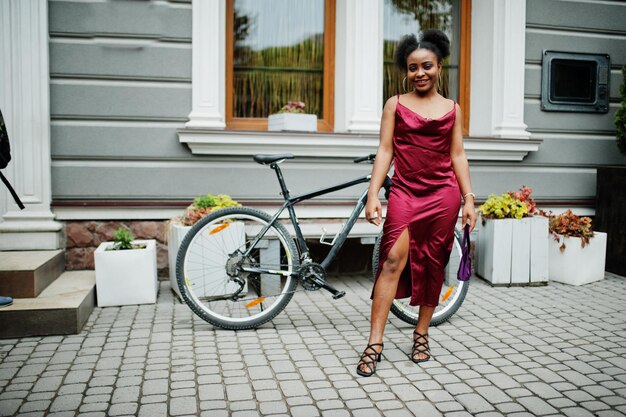 Portret van een mooie natuurlijke jonge Afrikaanse vrouw met afrohaar Zwart model in rode zijden jurk met mountainbike