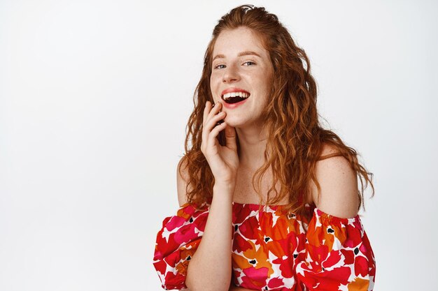 Portret van een mooie roodharige vrouw die lacht en glimlacht en die openhartige emoties toont die in jurk tegen een witte achtergrond poseren
