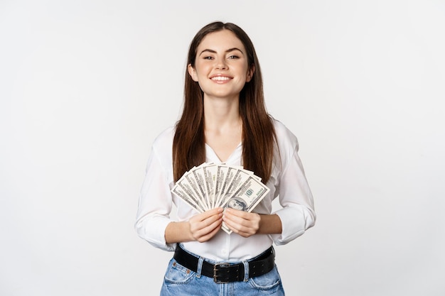 Portret van een mooie vrouw die geld contant geld aanhoudt en tevreden glimlacht terwijl ze op een witte achtergrond staat