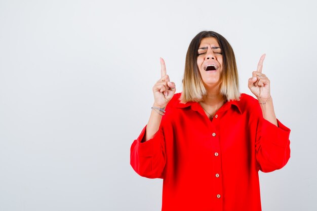 Portret van een mooie vrouw die naar boven wijst, de ogen gesloten houdt in een rode blouse en er een gelukzalig vooraanzicht uitziet