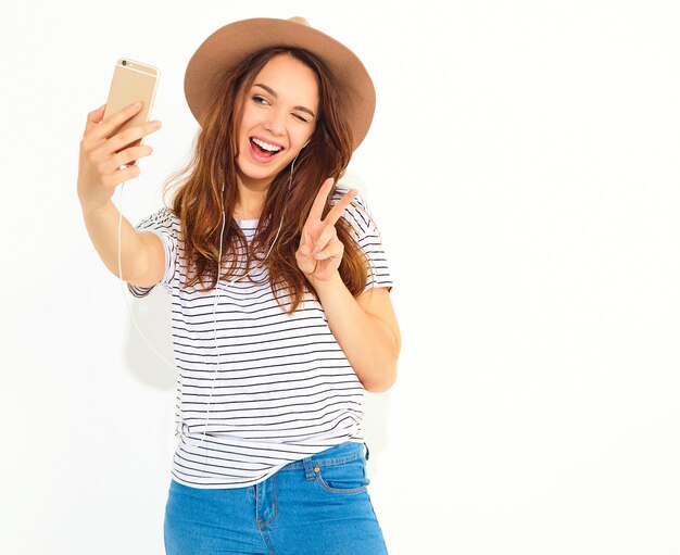 Portret van een mooie vrouw in de zomer hipster kleren die een selfie nemen op witte muur wordt geïsoleerd. Knipogen en vredesteken tonen