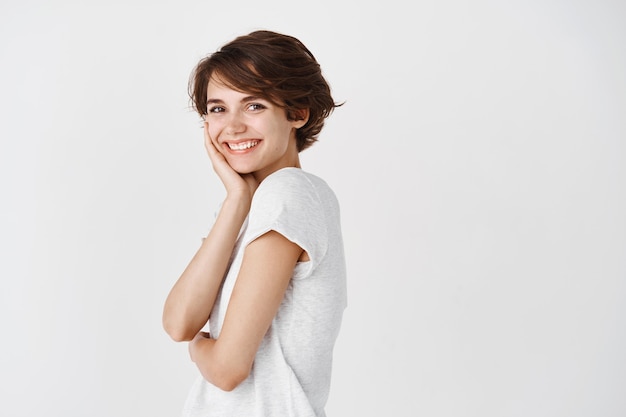 Portret van een natuurlijke jonge vrouw met kort haar, die een zuivere, schone huid aanraakt en glimlacht, staande tegen een witte muur