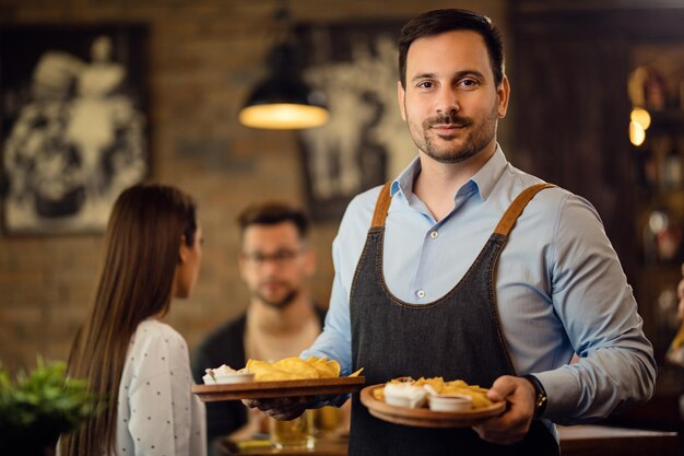 Portret van een ober die borden met eten vasthoudt en naar de camera kijkt terwijl hij in een pub werkt