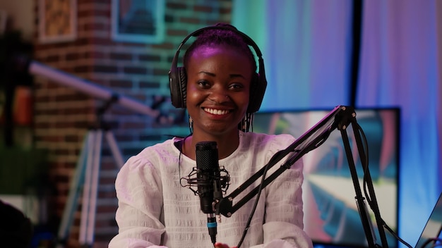 Portret van een online radiopresentator die zelfverzekerd naar de camera glimlacht terwijl hij live uitzendt met professionele apparatuur in de opnamestudio. Afro-Amerikaanse podcaster zit aan bureau met boomarmmicrofoon.