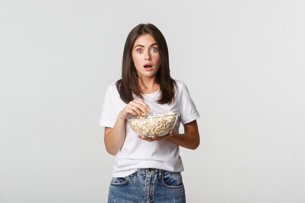 Portret van een opgewonden en verbaasde jonge vrouw die popcorn eet en films of tv-series kijkt.