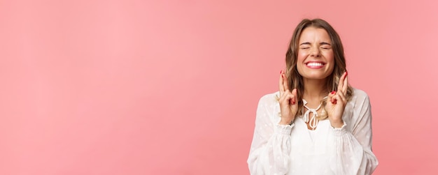 Portret van een opgewonden, hoopvol blond meisje dat een wens doet, gekruiste vingers voor geluk, sluit de ogen en glimlacht en steekt alle moeite in het bidden en smeekt om een droom die uitkomt, anticiperend op een roze achtergrond