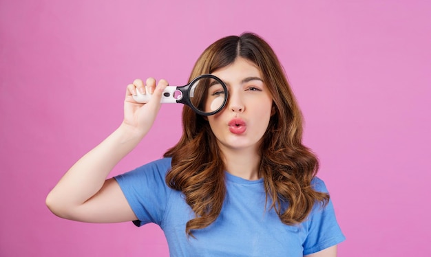 Portret van een opgewonden jonge vrouw die een casual t-shirt draagt en naar de camera kijkt door vergrootglas geïsoleerd over roze achtergrond