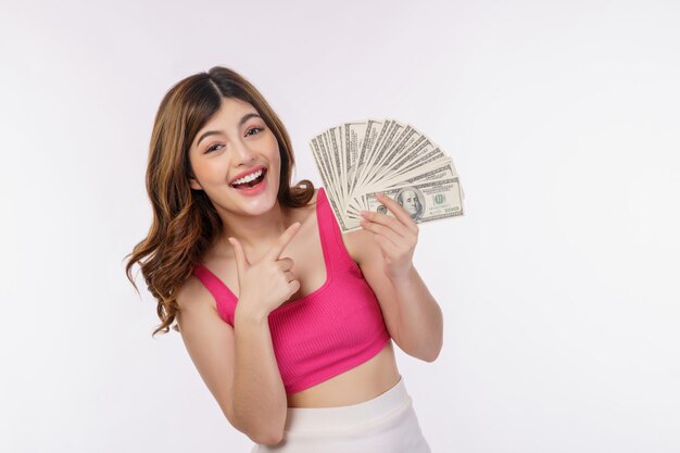 Portret van een opgewonden jonge vrouw die een stapel dollarsbankbiljetten vasthoudt en met de vinger naar geld wijst dat op een witte achtergrond wordt geïsoleerd