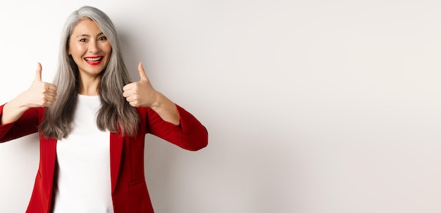 Portret van een oudere aziatische vrouw die een duim omhoog laat zien en een rode blazer draagt voor kantoorwerk