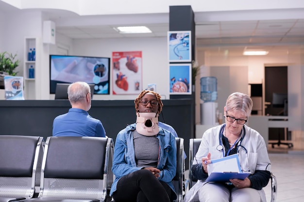 Portret van een patiënt met nekkraagschuim die in de wachtkamer in de kliniek zit en overleg pleegt met een senior arts. Vrouw die cervicale steun draagt na ongevalsverwonding die aan medic over gezondheidszorg spreekt.