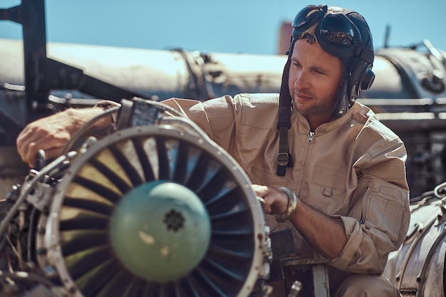 Gratis foto portret van een piloot-monteur in uniform en vliegende helm, die de ontmantelde vliegtuigturbine in een openluchtmuseum repareert.