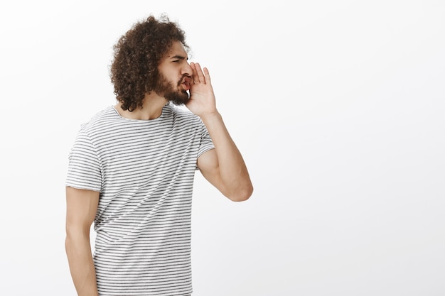 Portret van een pissige onbeleefde spaanse man, die naar rechts draait en de handpalm bij de mond houdt terwijl hij vloekende woorden hardop schreeuwt, fronsend, lastig gevallen en boos is