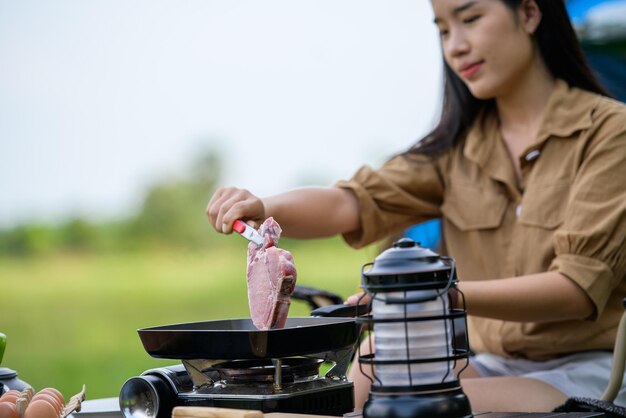 Portret van een portret van een gelukkige jonge aziatische vrouw die alleen kampeert met gegrild varkensvlees in de picknickpan en eten kookt terwijl ze op een stoel op de camping zit