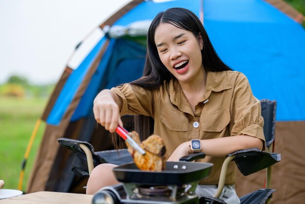 Portret van een portret van een gelukkige jonge aziatische vrouw die alleen kampeert met gegrild varkensvlees in de picknickpan en eten kookt terwijl ze op een stoel op de camping zit