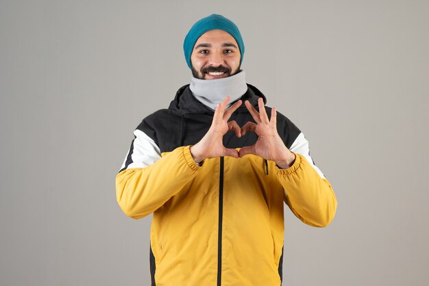 Portret van een positieve bebaarde man in warme kleren die staat en een hartsymbool doet met de handen.