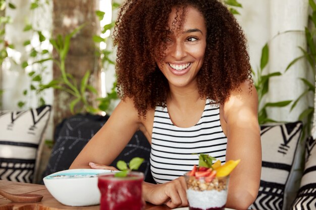 Portret van een positieve vrouw met een donkere huid heeft een positieve uitdrukking, kijkt gelukkig opzij, heeft een dromerige uitdrukking en denkt aan iets goeds, eet een smakelijk exotisch dessert, heeft een brede glimlach op het gezicht