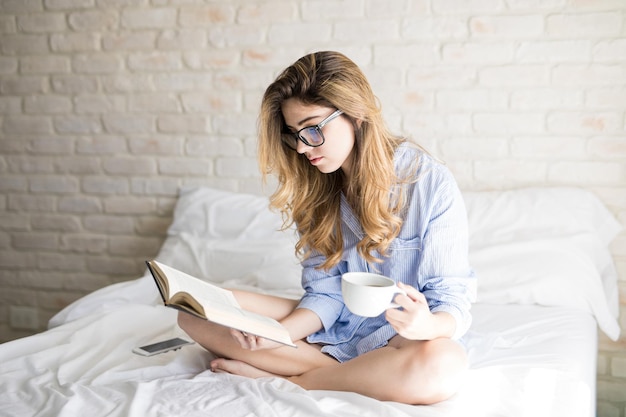 Gratis foto portret van een prachtig jong nerdy meisje met een bril die een boek leest en koffie drinkt in bed