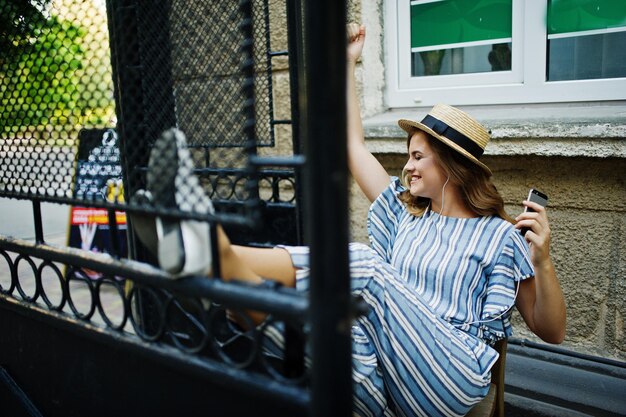 Portret van een prachtig model in gestreepte overall zittend op de stoel buiten en luisterend naar muziek in oortelefoons