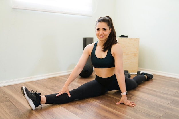 Portret van een prachtige brunette vrouw die een been splitst en glimlacht. Mooie sportieve vrouw die zich uitstrekt voor haar trainingsroutine thuis