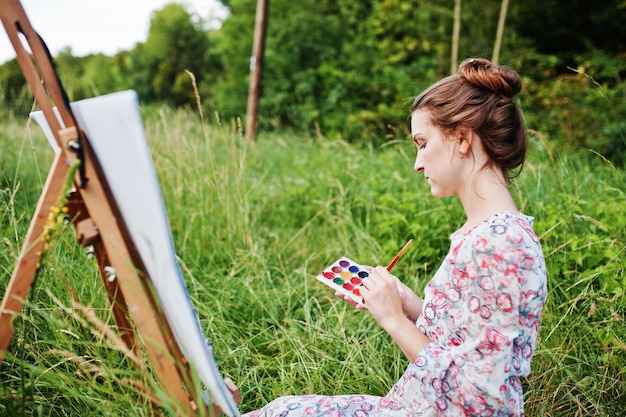 Portret van een prachtige gelukkige jonge vrouw in een mooie jurk zittend op het gras en schilderen op papier met aquarellen