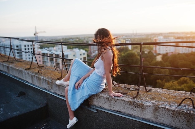 Portret van een prachtige jonge vrouw in wit t-shirt en blauwe rok zittend op de rand van het gebouw bij zonsondergang