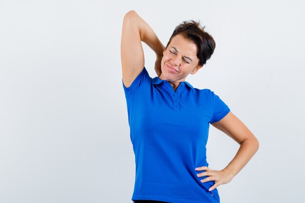 Portret van een rijpe vrouw die aan nekpijn in blauw t-shirt lijdt en moe vooraanzicht kijkt