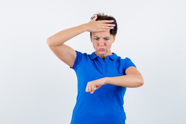 Portret van een rijpe vrouw die beweert horloge op haar pols in blauw t-shirt te bekijken en gealarmeerd vooraanzicht te kijken