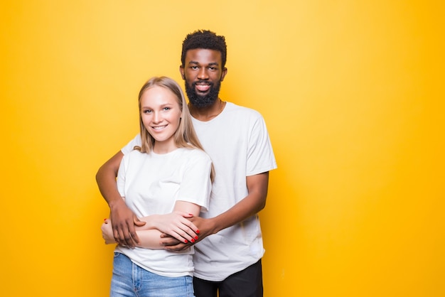 Portret van een romantisch multiraciaal paar dat elkaar omarmt, samen poseert en glimlacht, over een gele muur staat