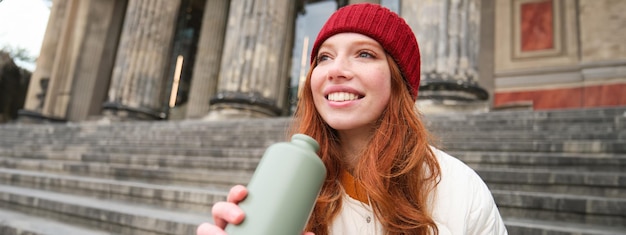 Gratis foto portret van een roodharige vrouw die drinkt uit een thermosfles die op de straattrap zit en geniet van een warme drank