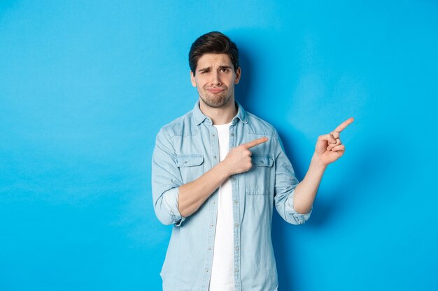 Portret van een sceptische volwassen man die met de vingers naar rechts wijst en grijnst, teleurstelling en twijfel uitdrukt, staande tegen een blauwe achtergrond