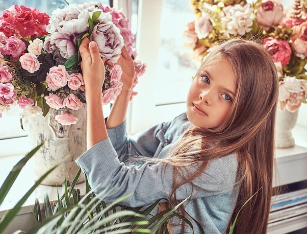 Portret van een schattig klein meisje met lang bruin haar en doordringende blik in een stijlvolle jurk, poserend met bloemen tegen het raam thuis.