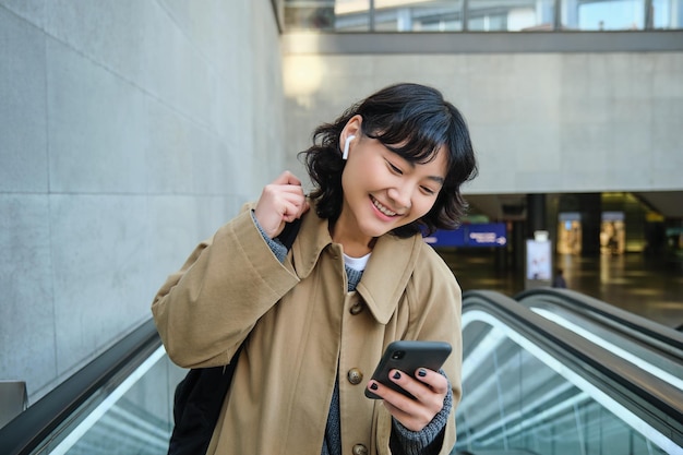 Portret van een schattig Koreaans meisje in een trenchcoat die de roltrap opgaat, houdt mobiele telefoon vast en glimlacht tevreden