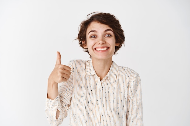 Portret van een schattig ondersteunend meisje dat duimen opsteekt en trots glimlacht, je prees, goed gedaan, uitstekend gebaar laat zien, tevreden op de witte muur staat