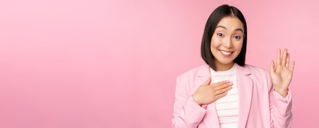Portret van een schattige aziatische zakenvrouw die de hand opsteekt, stelt zichzelf voor op kantoor en lacht koket over een roze achtergrond in een pak