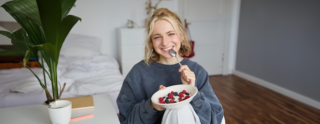 Portret van een schattige glimlachende blonde vrouw die ontbijt eet in haar slaapkamer en naar een schaal kijkt die een camera vasthoudt