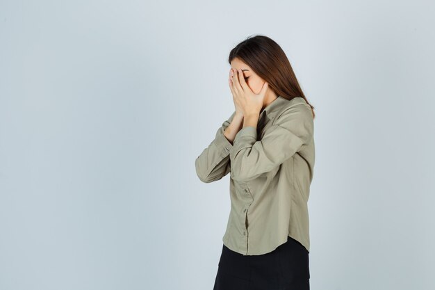 Portret van een schattige jonge vrouw die het gezicht bedekt met de handen in het hemd, de rok en een depressief vooraanzicht kijkt