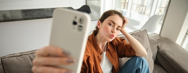 Gratis foto portret van een schattige jonge vrouw, een modern meisje dat selfies maakt op een smartphone-app en op de bank poseert