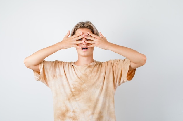 Gratis foto portret van een schattige tienerjongen die door de vingers in een t-shirt gluurt en zich afvraagt vooraanzicht