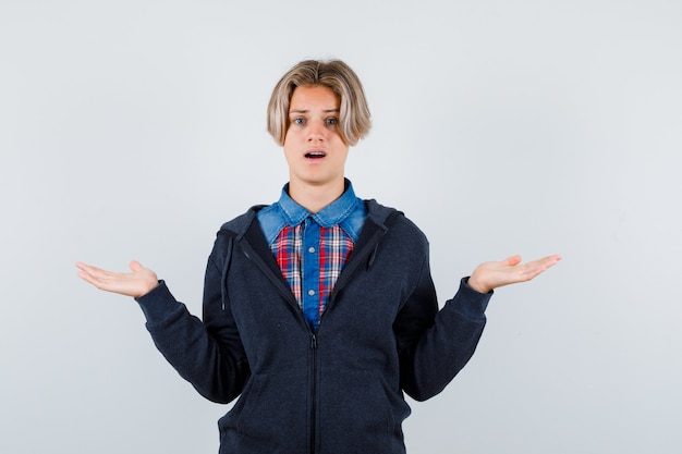 Portret van een schattige tienerjongen met een hulpeloos gebaar in shirt, hoodie en een verbaasd vooraanzicht