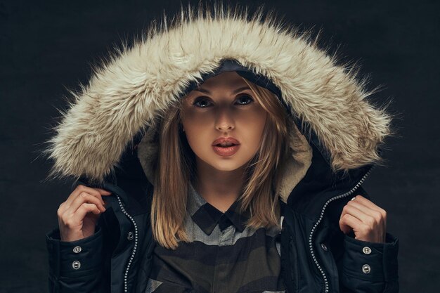 Portret van een sensueel blond meisje in een winterjas en bontkap, staande in een studio. Geïsoleerd op een donkere achtergrond.